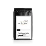 Adiramo 6 Bean Blend 12oz black coffee bag with white label standing next to a steaming mug and fresh roasted whole coffee beans