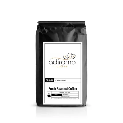 Adiramo 6 Bean Blend 12oz black coffee bag with white label standing next to a steaming mug and fresh roasted whole coffee beans